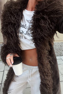 Dark brown faux fur coat with shaggy front trim and cuffs, styled with grey sweatpants on a city street