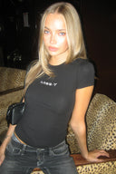 Black tight-fit baby tee with silver Miss Y logo and crew neck, styled with grey jeans, in a dimly lit lounge