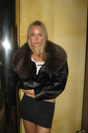 Black faux leather baggy jacket with brown faux fur collar, styled with a black mini skirt in a tiled hallway