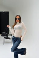 Off-white sheer tight-fit turtleneck body with long sleeves, styled with blue flared jeans, in a white photo studio