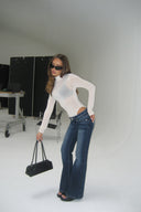 Off-white sheer tight-fit turtleneck body with long sleeves, styled with blue flared jeans, side view in a white photo studio