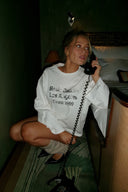 Off-white loose-fit crew neck top with long sleeves and text print, styled with black heels in a hotel corridor