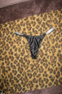 Black satin low-waist thong with rhinestone-studded elastic side straps, laid flat on a leopard print rug